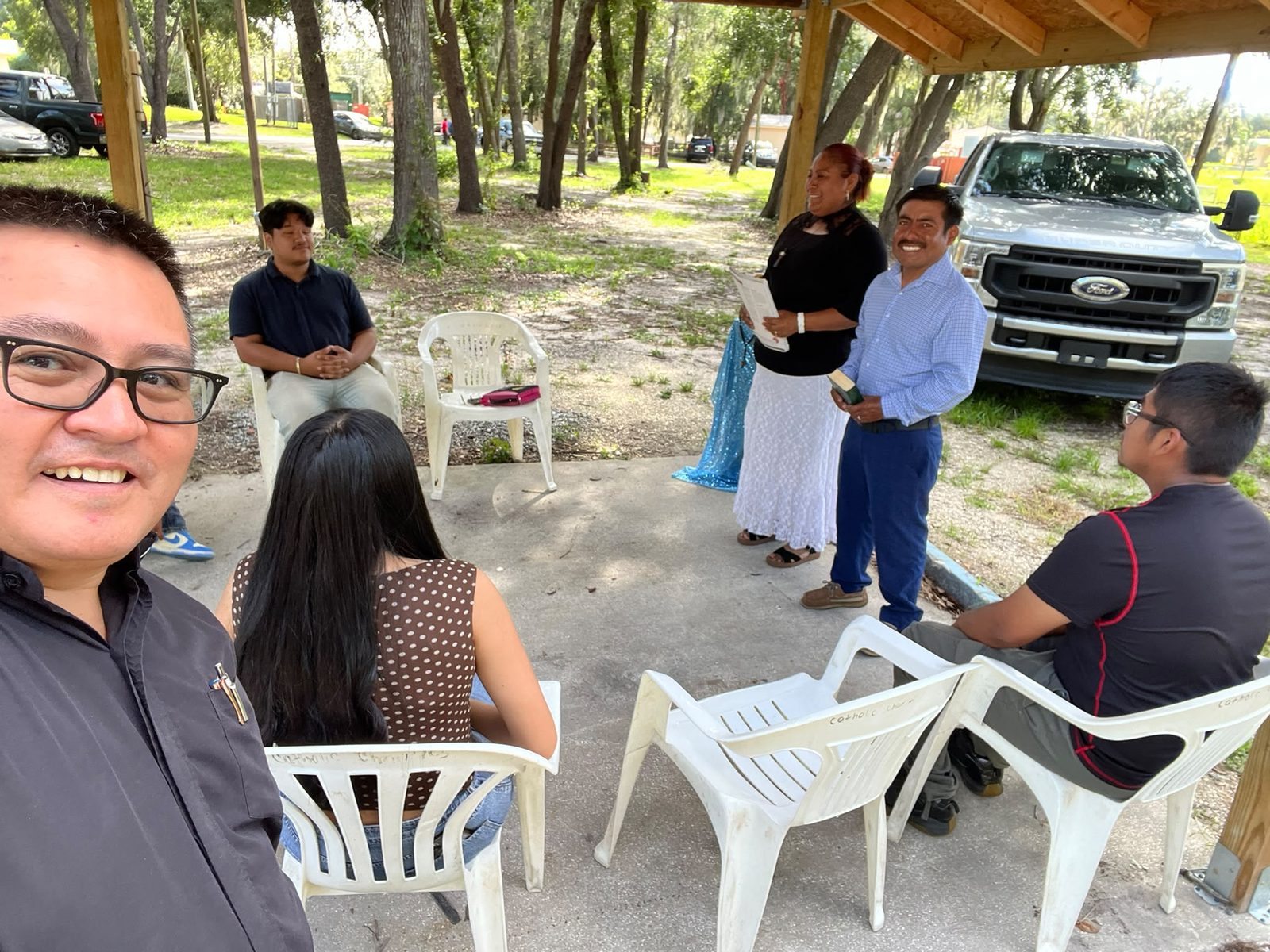 A small group gathered under the Mission courtyard shelter, with the priest in the foreground