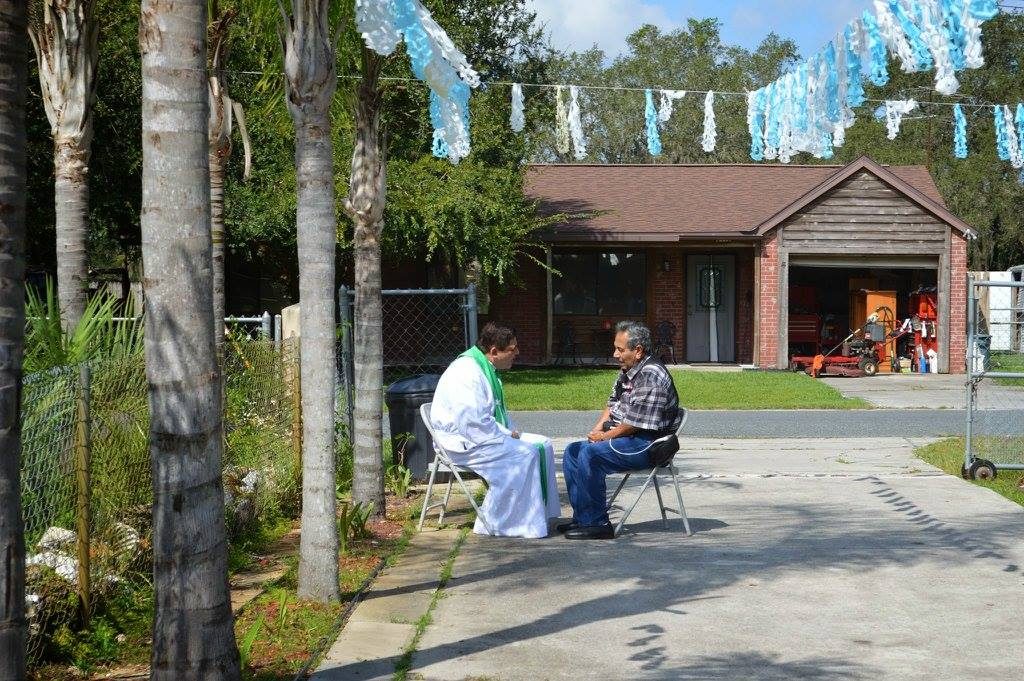 A priest in a green stole hears a man's confession, outdoors in front of a house